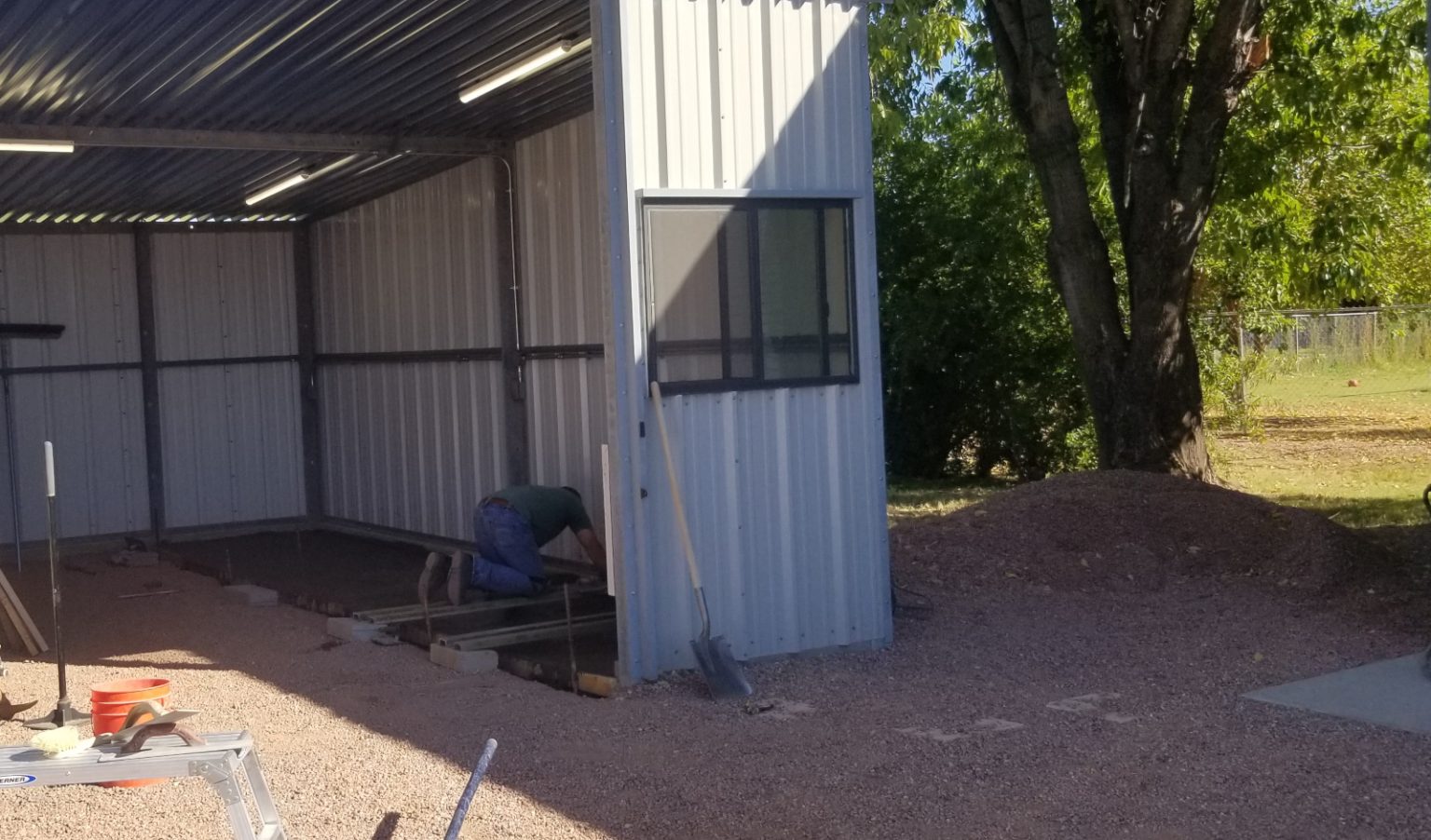 Metal carport shed in Gilbert, AZ. Man on his knees working a freshly poured concrete slab. Trees, sunny day.