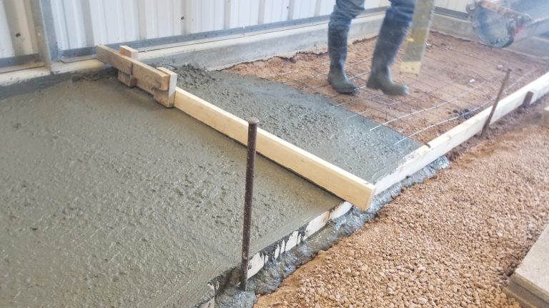 A man's legs are visible, he is wearing concrete boots, fresh concrete has just been poured, a screed board is sitting on top of the concrete.