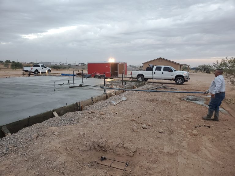 Concrete slab foundation freshly poured at a residential construction site in a desert area, with workers finishing the surface and trucks parked nearby.