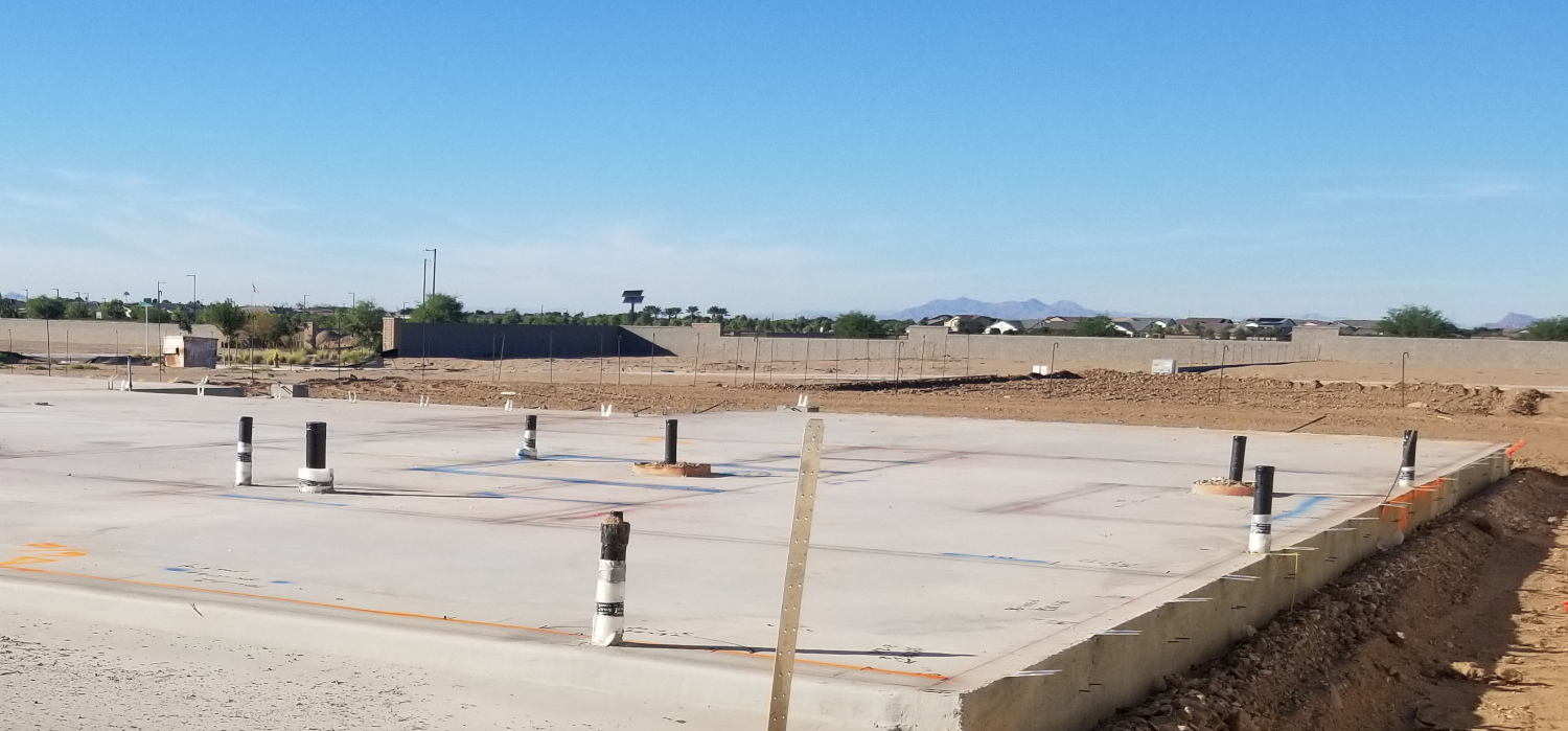 Construction site in Queen Creek, AZ. a flat concrete house pad with plumbing sticking out of the slab, ready for a new home to be built on top of it. Blue sky, construction field, and neighborhood in the background.
