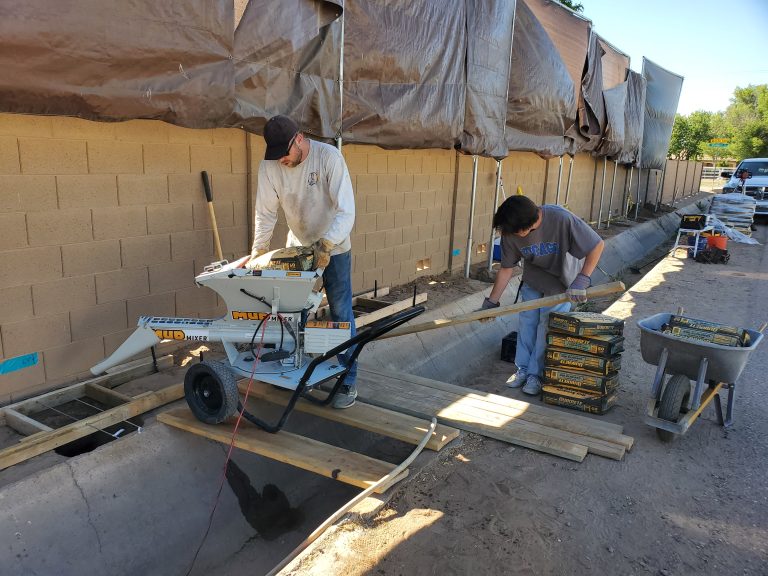 Two men pouring concrete with a small mud mixer while straddling over an irrigation canal.