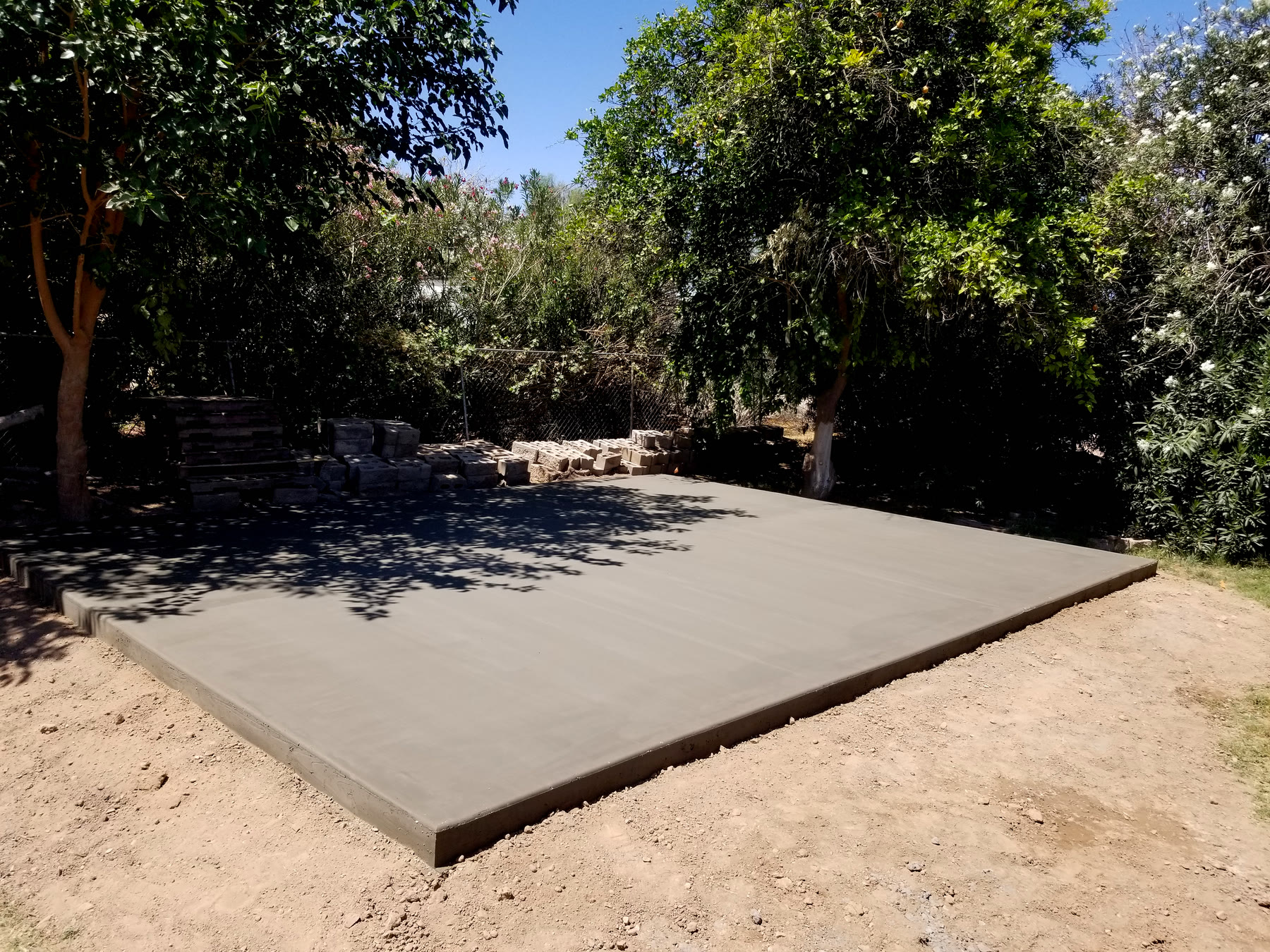 Wide view of a finished concrete shed pad in a tree-lined Gilbert backyard